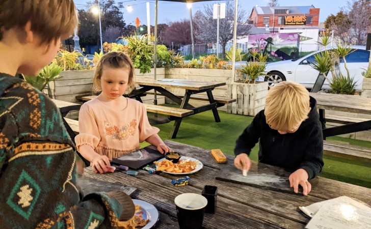 three children at wooden al fresco table in lush garden draw on blackbords and enjoy a kids meal
