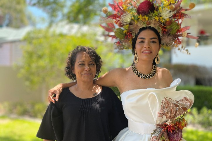 mother stands with her daughter who's wearing a white hand painted dress and floral head dress
