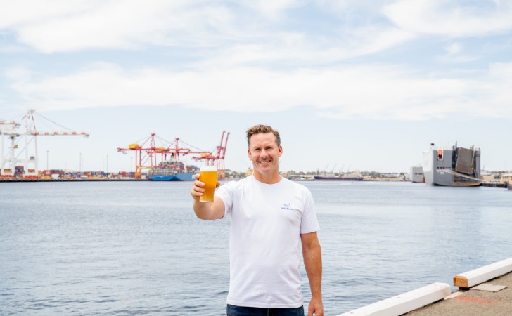 person holds beer, standing at waterfront with huge ship and port cranes behind them