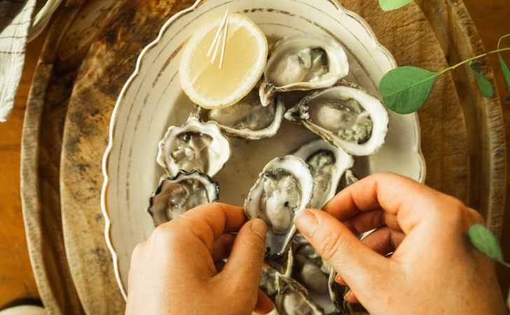 hands shucking fresh oysters on a plate with lemon and green foliage