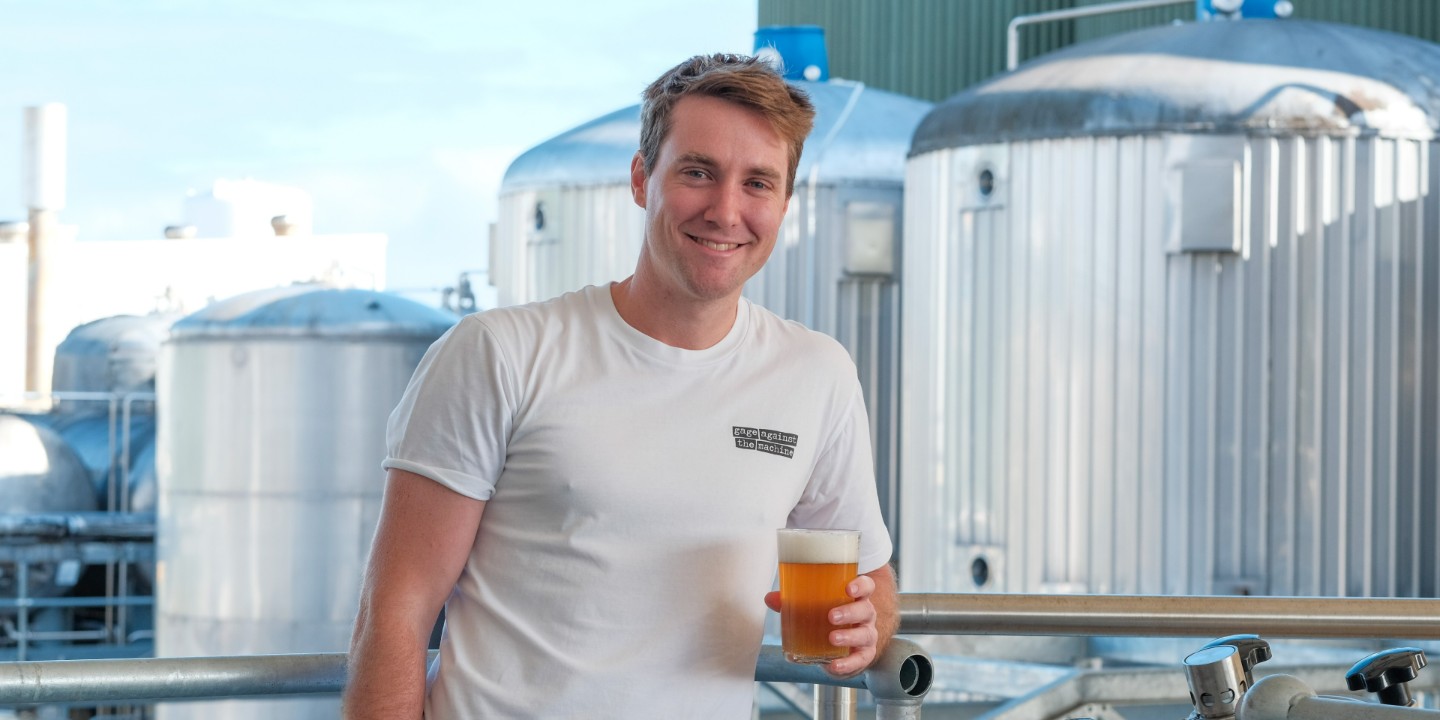 cam parker wears white 'gage against the machine' shirt and holds a pint, standing in front of brewery