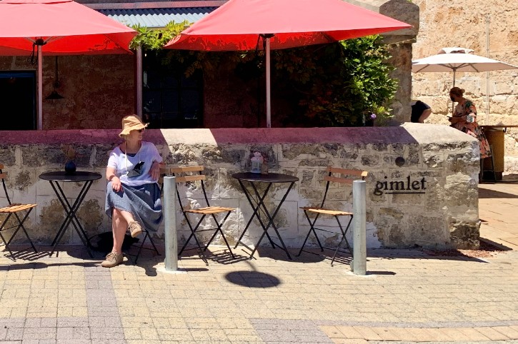 person wearing sunglasses and hat sits at a small table in front of heritage limestone brick wall which says 'Gimlet'