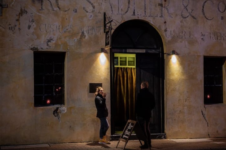 two people in dark clothing stand outside curtained door of quirky heritage building 