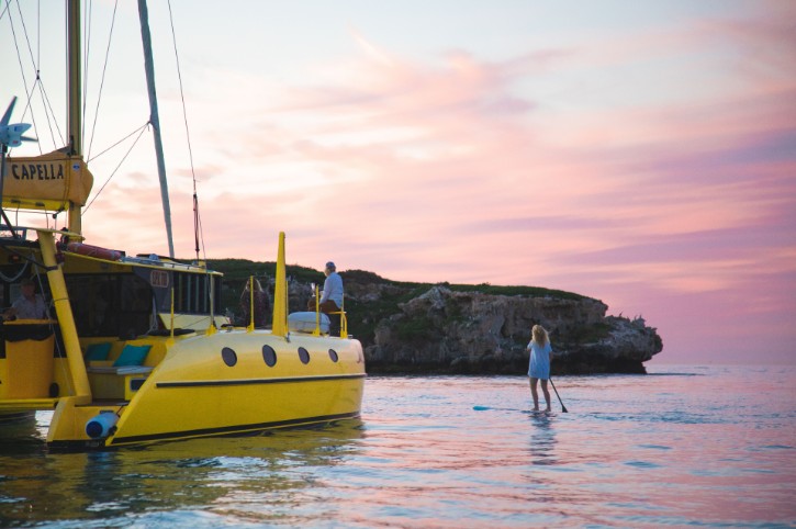 person stand on yellow catamaran watching their partner on stand up paddle board under pink sunset