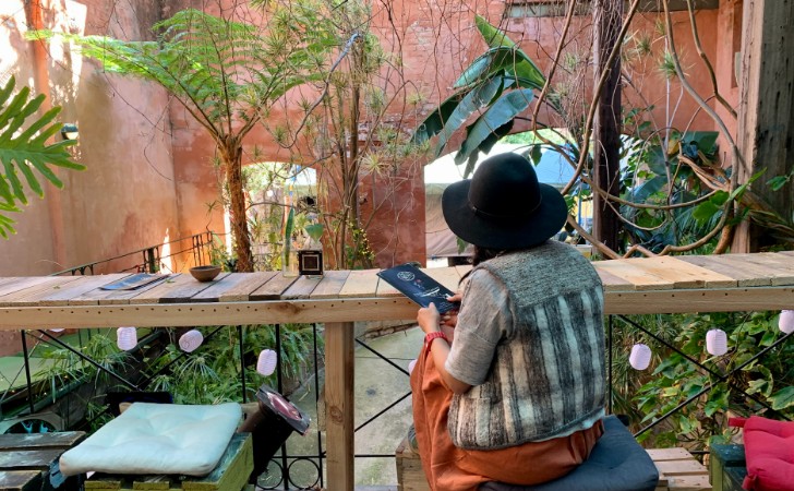 person wearning black hat sits at wooden cafe table facing lush green gardens