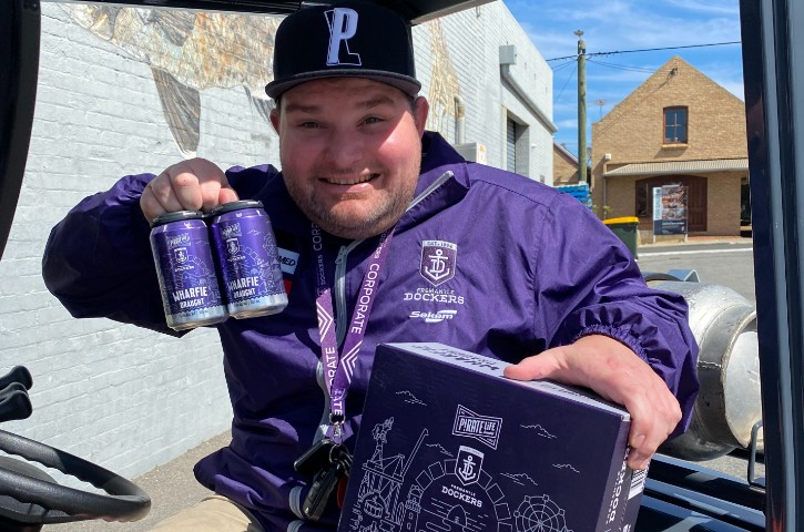 person wearing purple Fremantle Dockers jacket and black Pirate Life Brewing hat holds tins of Wharfie Draught beer