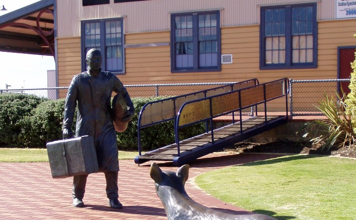 bronze statue of a person with suitcase returning to dog in front of E Shed Markets, painted in heritage mustard
