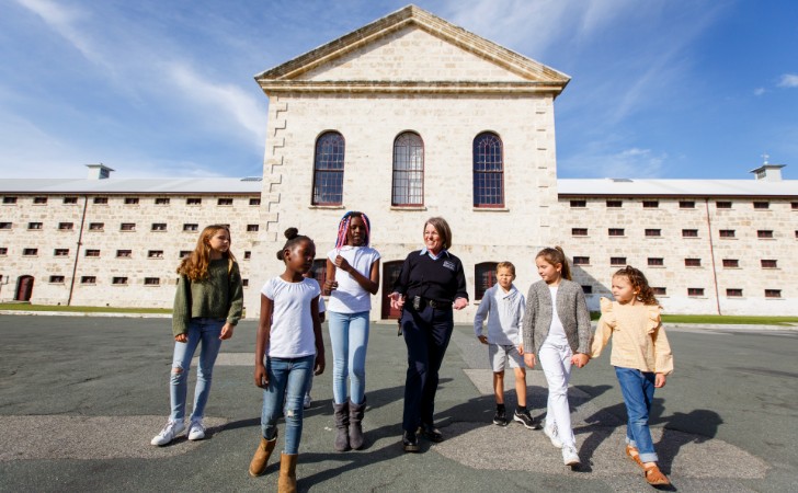 a group of children tour with prison guard in front of limestone heritage building and clear blue sky