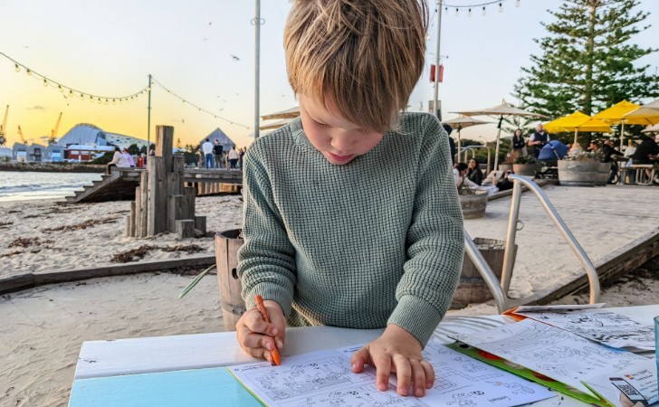 child enjoys colouring in, with white sandy beach, fairy lights and sunset behind them