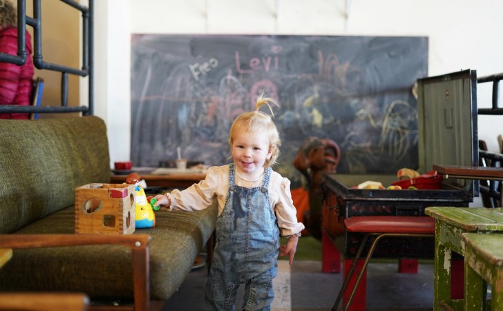 small smiling child in overalls stands in cafe playground with giant blackboard and retro green couches