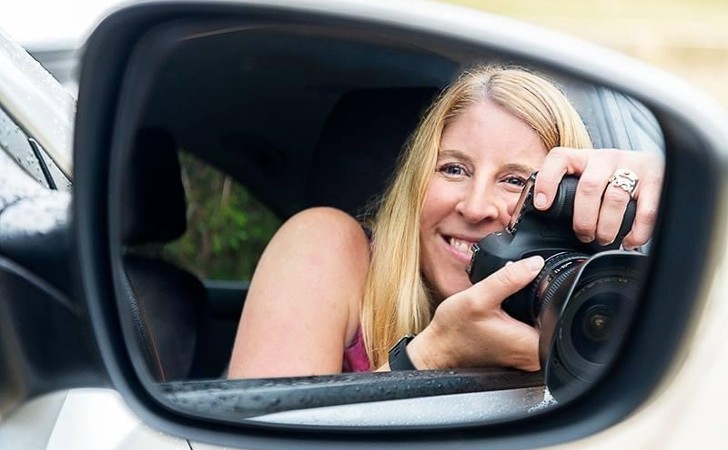 photographer smiles while taking photo in rear view mirror with a camera