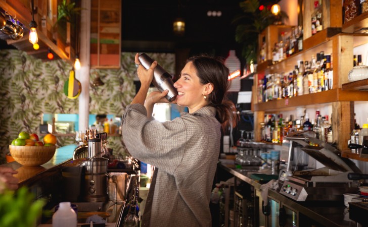 smiling person shakes a cocktail in a kooky, well-stocked bar
