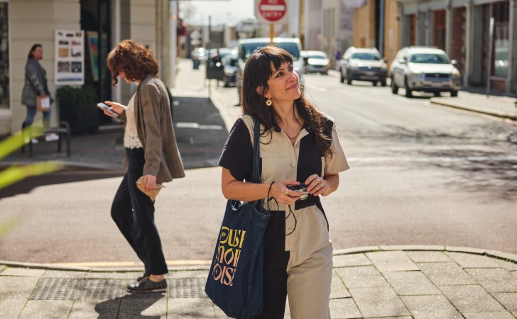 person wearing cool jump suit and carrying shoulder bag holds camera while looking up at heritage buildings in car-lined street