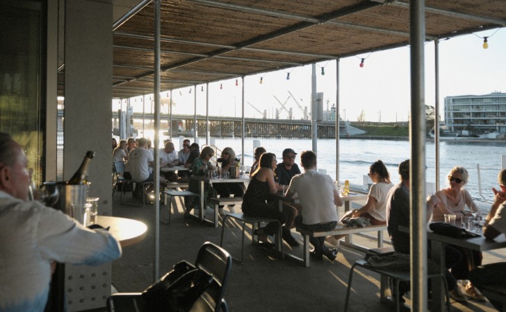 people eating and drinking at restaurant looking out over still water, bridge, cranes and sunshine