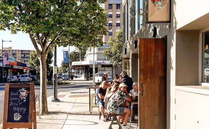 people sit out at tables in tree lined street with sign saying "Don't worry, beer happy"