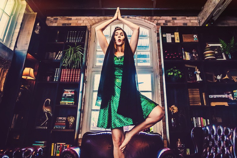person wearing sparkly green dress and black veil stands on one leg on a vintage leather chair in a library