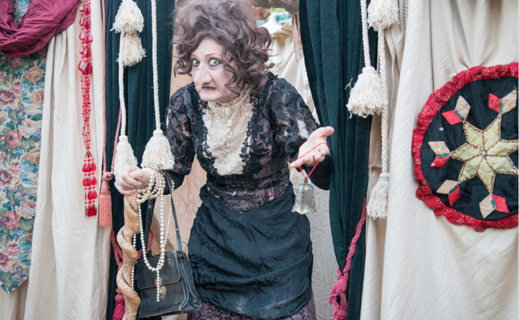 Fremantle International Street Arts Festival artist on stage in front of cream drapes, dressed up with fake nose in old fashioned black lacey dress 