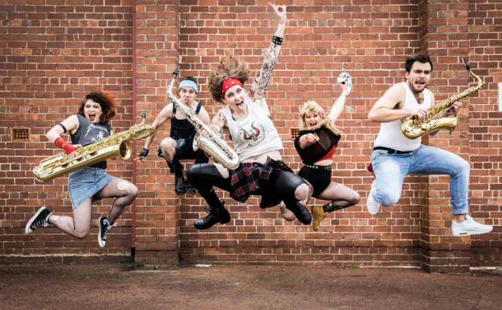 Fremantle International Street Arts Festival artists Sax Rockers - five musicians jumping off the ground holding saxophones in front of brick wall 