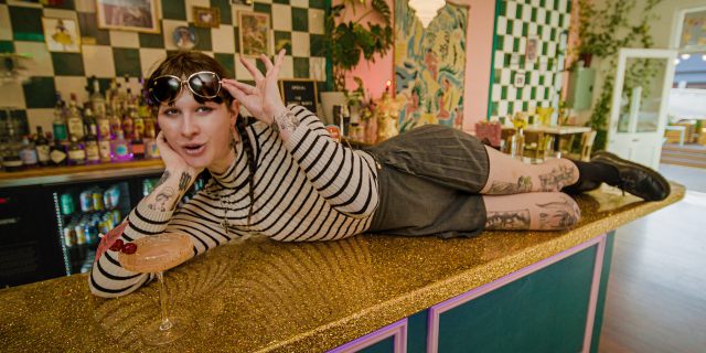 person in striped shirt lounges on gold sparkly bar with fruity cocktail