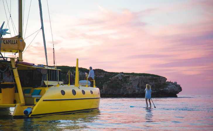 person stand up paddle boarding in front of luxury yellow catamaran beneath pink sunset