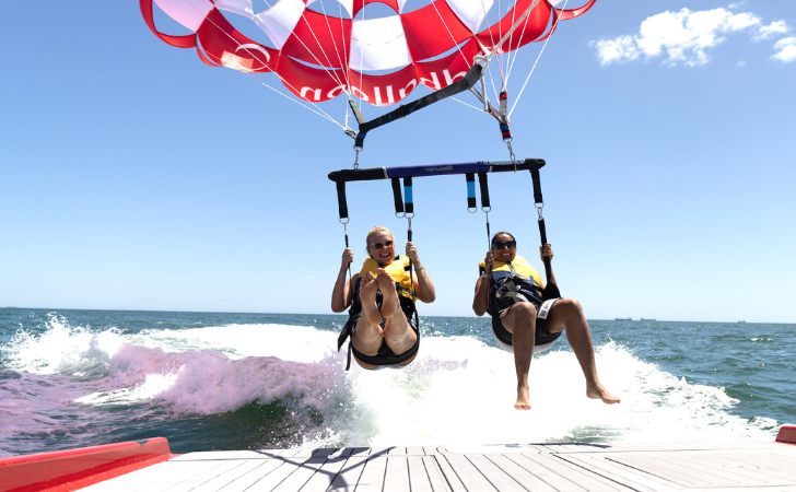 two people parasail over turquoise waters with red and yellow striped parachute