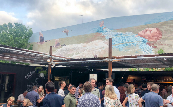 Huge mural of a man floating with geese above him is above a bustling small bar beer garden