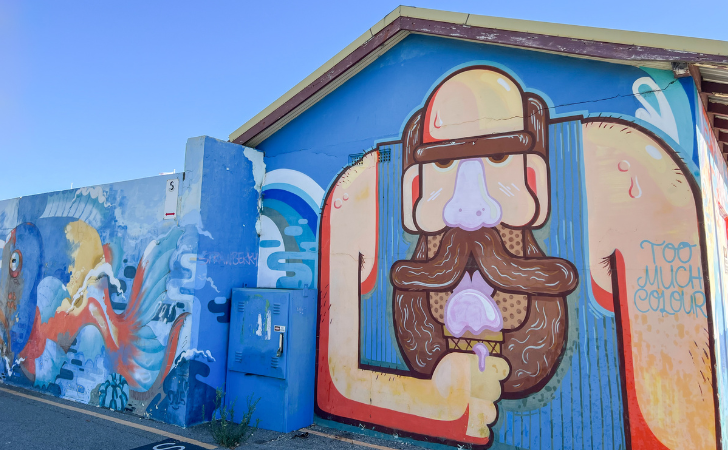 painting of bearded, singeted person licking a pink ice cream on a blue building
