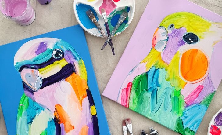 Two paintings of colour parrots on a table with paint brushes and pots scattered around.