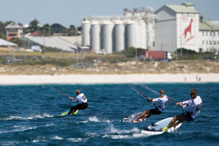 Three kitesurfers on the ocean with a flour mill in the background.