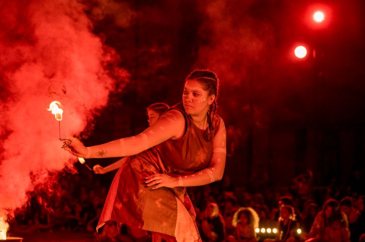 A young indigenous Australian woman dancing in the dark with red lights shining on her, holding a fire stick.