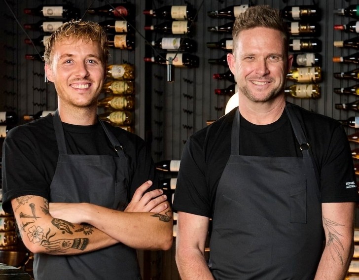 Two chefs in black t-shirts and black aprons standing in front of a wine cabinet, smiling at the camera.