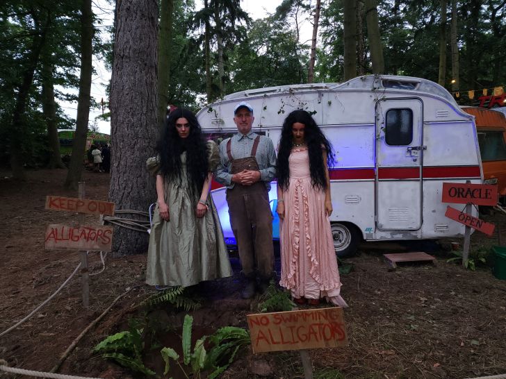 A man and two women stand in front of a beaten up trailer in the woods, looking menacingly into the camera.