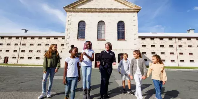 a group of children tour with prison guard in front of limestone heritage building and clear blue sky