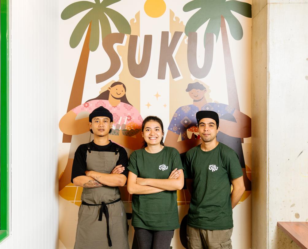 Three staff members from Suku standing in front of the a wall painted with palm trees 