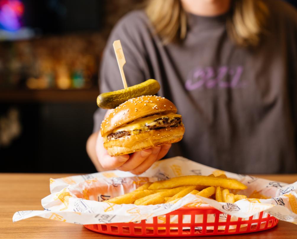 Person wearing a grey tshirt holding a burger with a pickle on top. Red basket of chips lined with Varsity paper. 