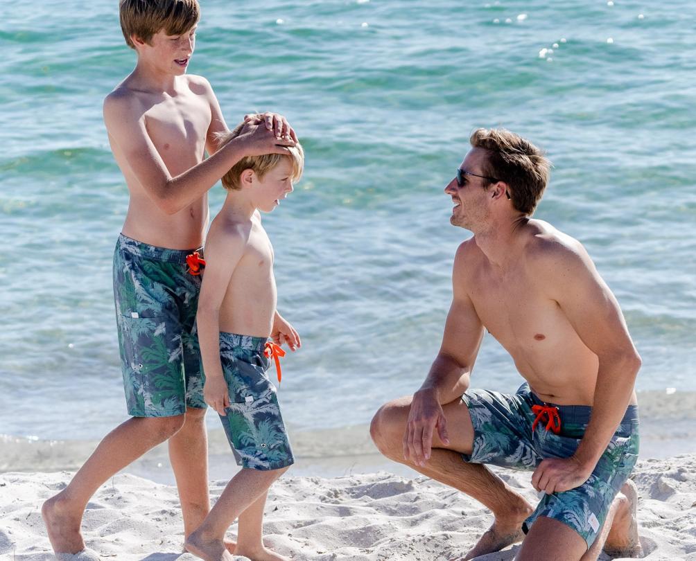 Dad on beach smiling at with two young boys wearing matching board shorts 