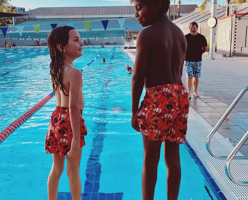 two children wearing board shorts standing at the end of a swimming pool. 