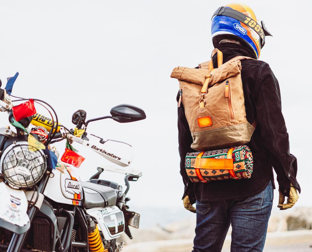 person wearing black jacket, motorcycle helmet and brown backpack stands next to motorbike