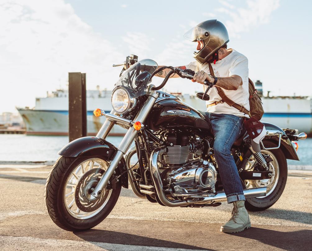 person wearing white t-shirt, jeans and helmet sits on motorbike in front of ocean with big ship