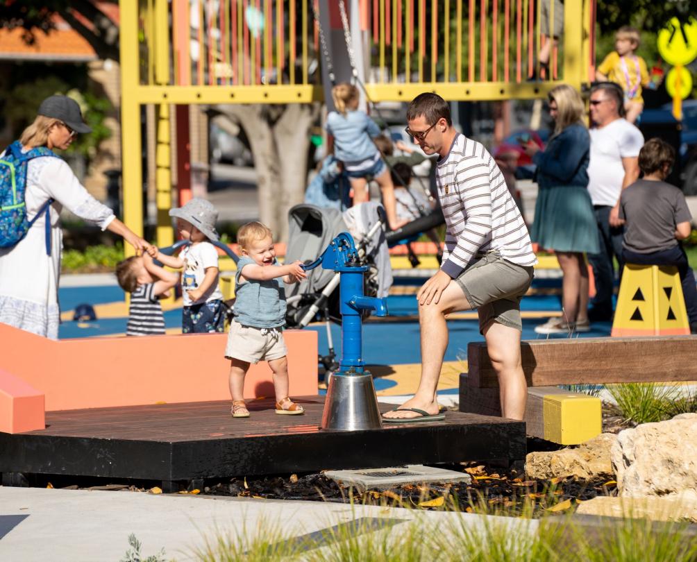 Child playing with water pump, watched by parent at Walyalup Koort playground