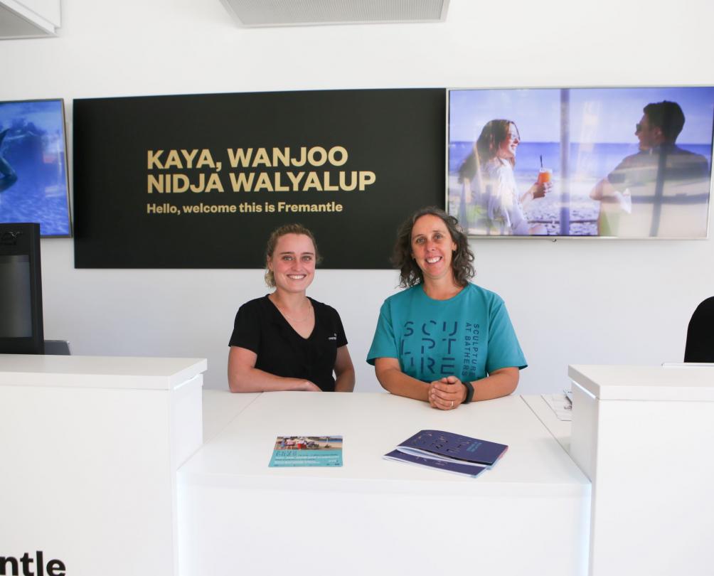 Fremantle Visitor Centre staff two women stand behind counter smiling. Black sign in the background with text Kaya, Wanjoo Nidja Walyalup