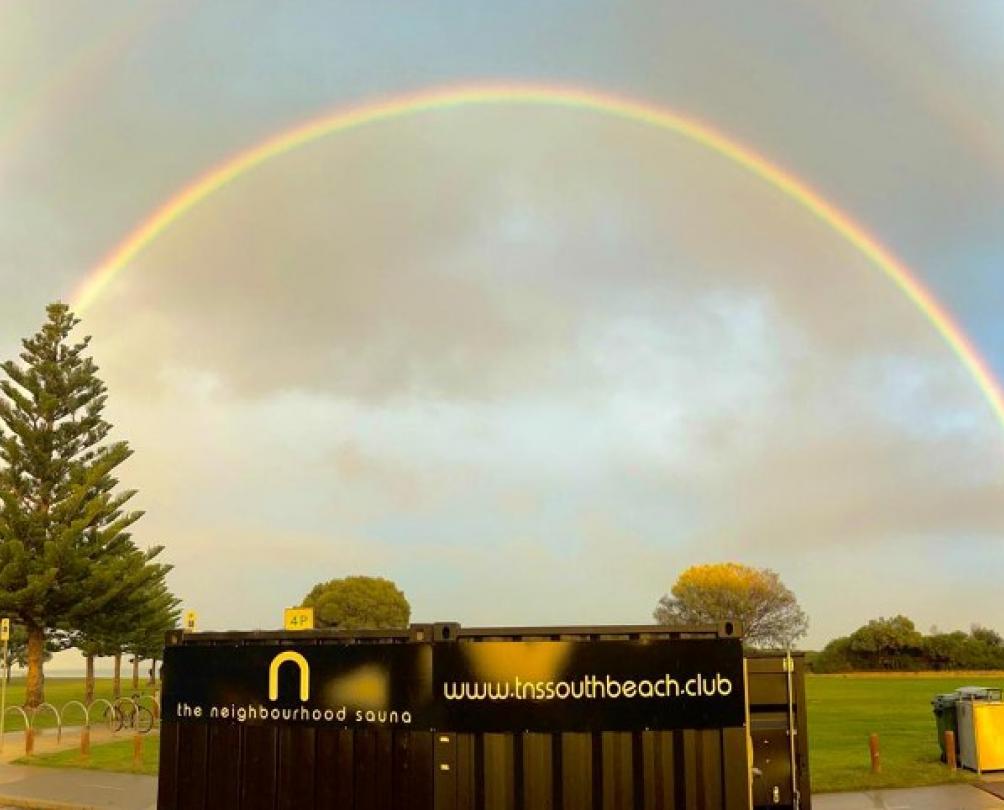 rainbow arches in blue sky over a beach front sauna near grass and tall Norfolk pine trees