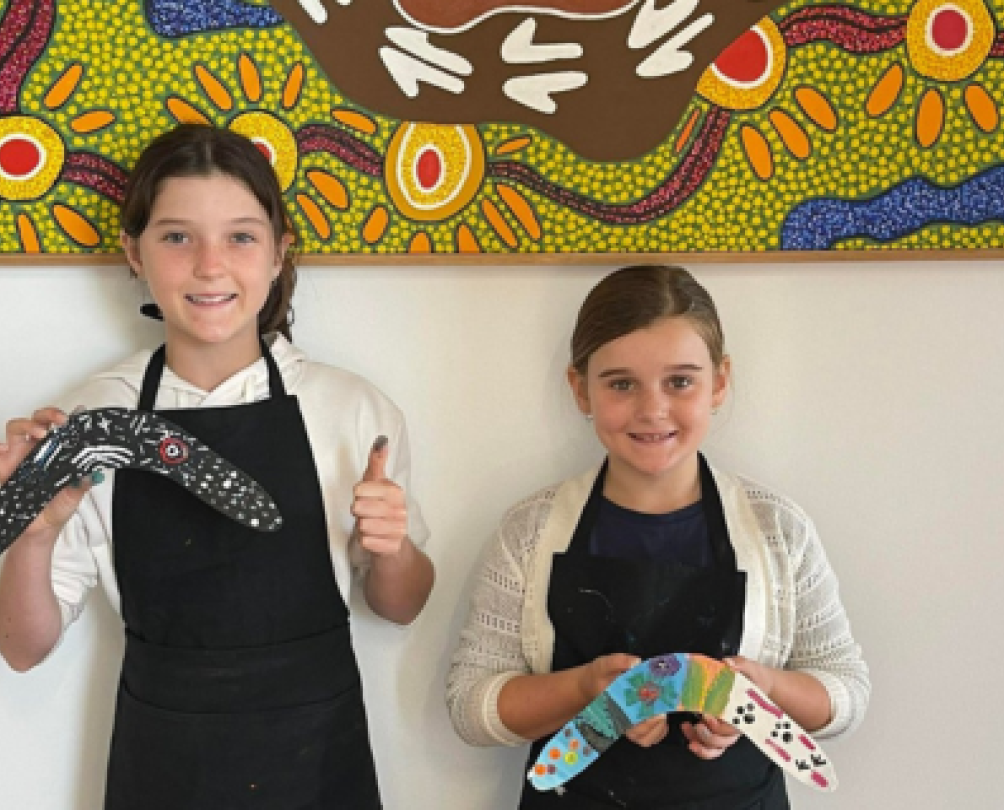 children hold hand painted boomerangs in front of colourful aboriginal art