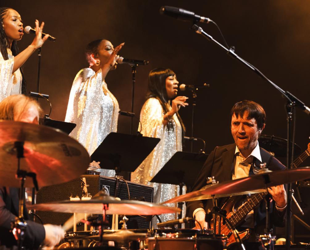 Three women dressed in sparkling silver gowns sing on a screen behind a band.