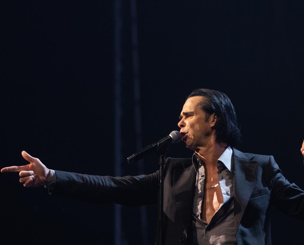 A man in a black suit and grey shirt half open sings into a microphone.
