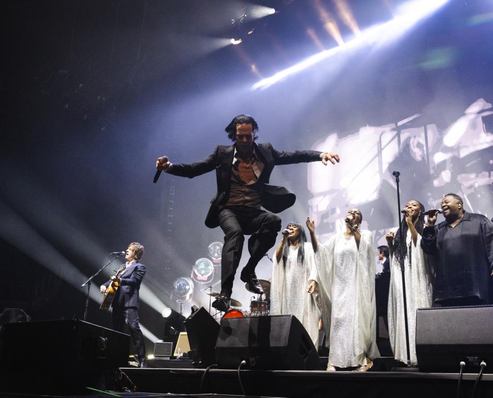 A musician dressed in a black suit jumps off a stage with a choir behind him.