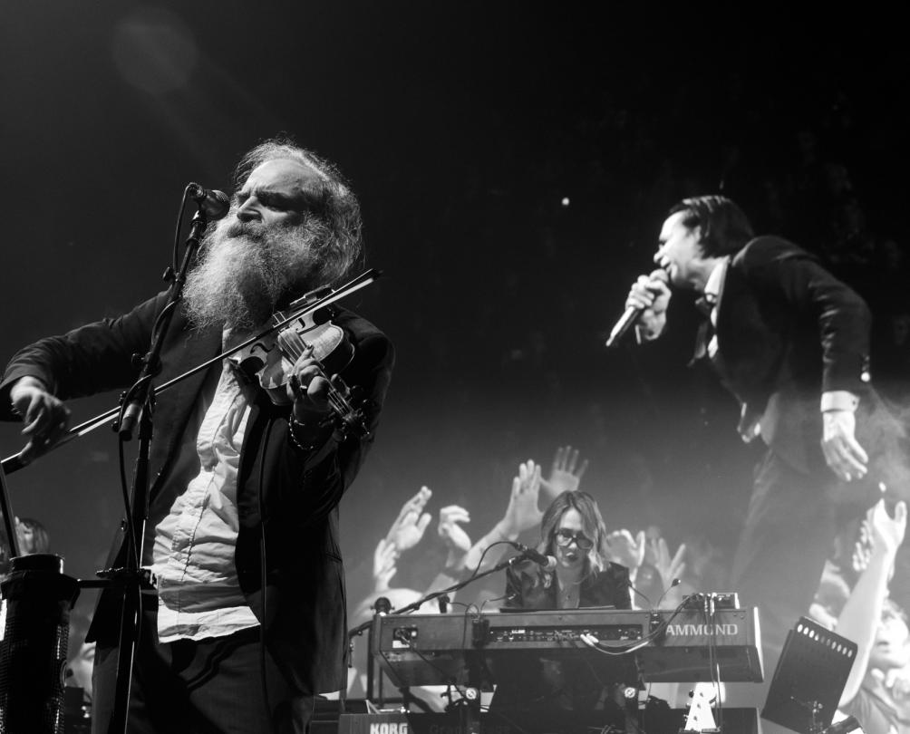 A man with a full beard plays the violin while a man in a black suit sings on a screen behind him.