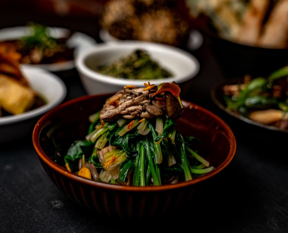 A close up of a bowl of Asian-style greens with several other dishes behind.