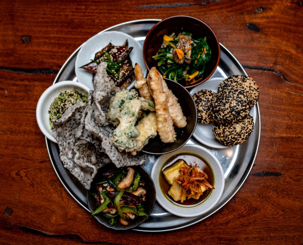 A silver platter on a wooden table filled with small Asian dishes to share.