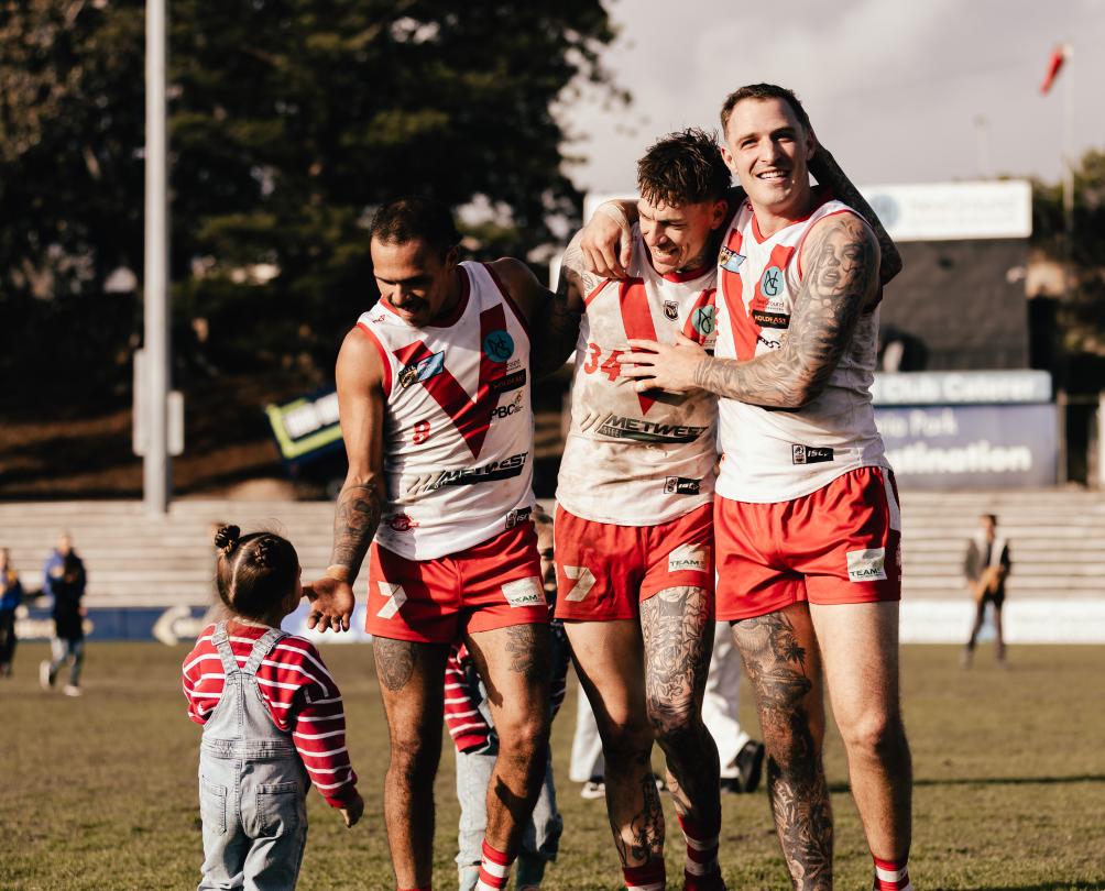 Three men in red and white football jerseys celebrate after a win as a small child dressed in red and white runs towards them.